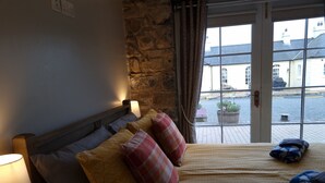 A bedroom with a stone wall, a bed with patterned pillows, a bedside lamp, and a view of the street through a window. - Cobbles Cottage - 2 Bedroom Stone Built Cottage (Ballynahinch)