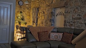 A living room with a stone wall, a black leather sofa, a woven coffee table, and a dining area with a chandelier. - Cobbles Cottage - 2 Bedroom Stone Built Cottage (Ballynahinch)