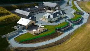 An aerial view of a large, historic building complex with multiple structures, including a central building with a large roof and several smaller buildings. There is a paved driveway leading to the entrance, and the area is surrounded by grassy fields. - Cobbles Cottage - 2 Bedroom Stone Built Cottage (Ballynahinch)