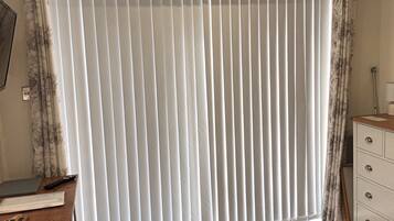 A room with a large window covered by vertical blinds, a wooden desk with a laptop and remote control, and a white dresser with drawers.