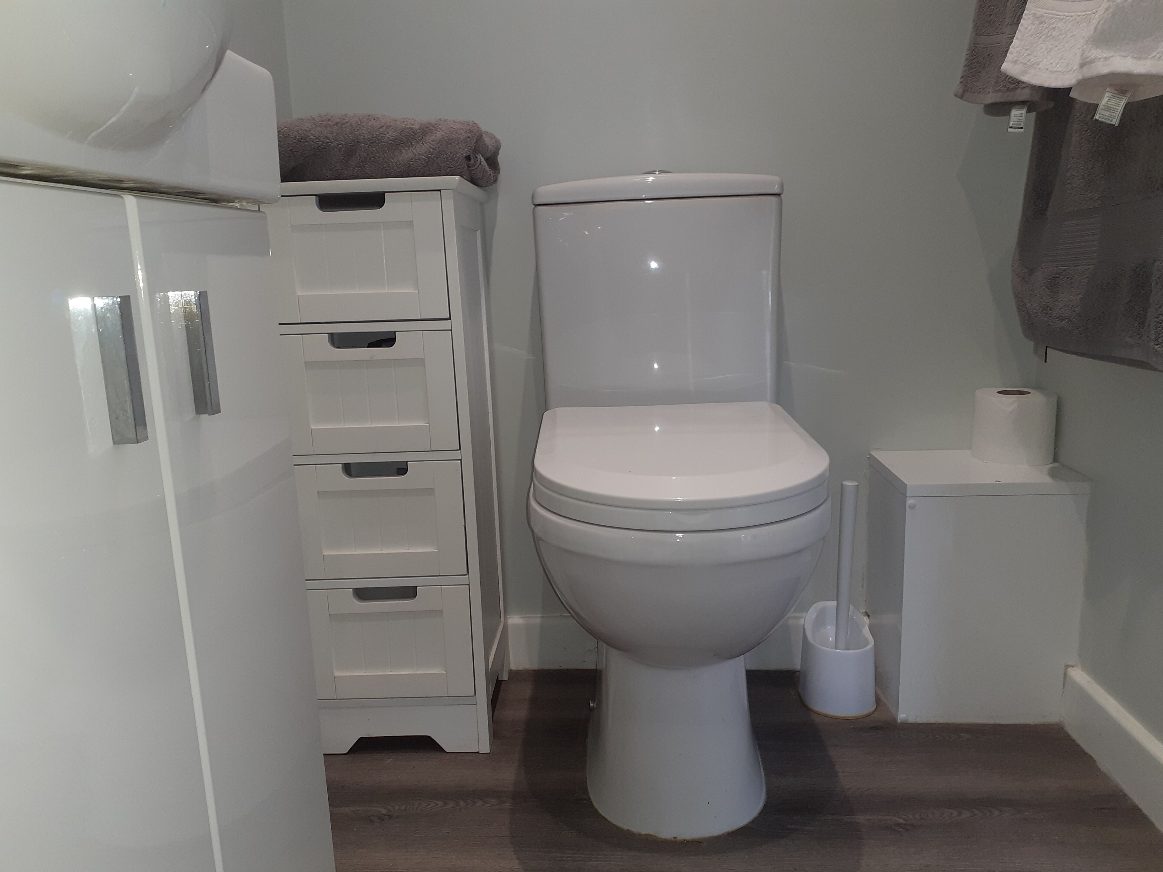 A small, clean bathroom with a white toilet, a white cabinet with drawers, and a roll of toilet paper on a small white stand.