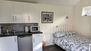 Eine kompakte Küche mit Mikrowelle, Wasserkocher und Spüle. Ein Bett mit floralem Überwurf und ein gerahmtes Bild an der Wand.