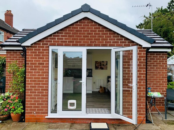 A brick house with a gabled roof and a sliding glass door leading to an interior with a kitchen and a living area. - Rose View - Modern Detached Bungalow (Sale)