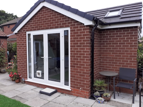 A brick house with a white-framed sliding glass door, a small outdoor seating area with a table and chairs, and potted plants around the entrance.