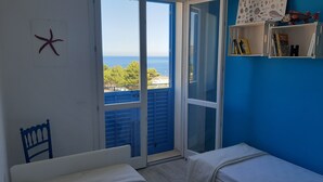 A room with a bed, a chair, a starfish decoration, a shelf with books and a fish model, and a view of the sea through a large window.