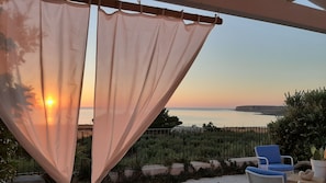 Una terraza con vistas a la puesta de sol sobre el mar, una silla azul y una planta en maceta.
