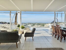 A covered outdoor seating area with wicker furniture and a view of the city and sea.
