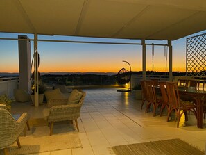 Eine Terrasse im Freien mit Korbmöbeln, einem Hängesessel und Blick auf den Sonnenuntergang über den Bergen.