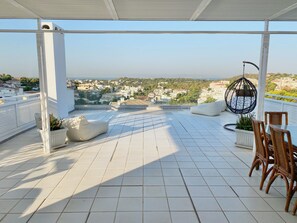 Eine Dachterrasse mit Fliesenboden, weißen Säulen, einem Hängestuhl und Blick auf eine Stadtlandschaft.