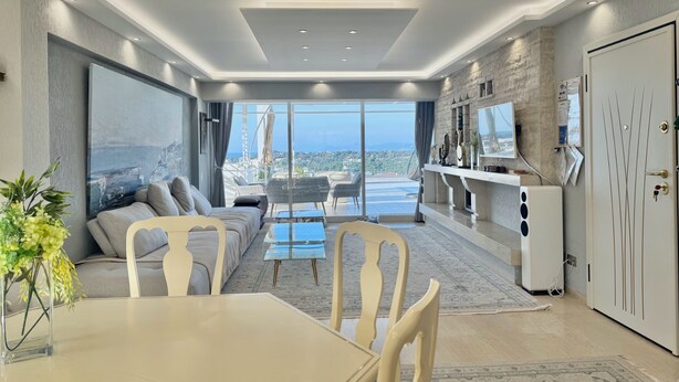 A modern living room with a sofa, dining table, and chairs, a large window with a city view, and a kitchen area with a refrigerator and counter. - Villasonboard 3 Bedroom Penthouse Vouliagmeni (Vari-Voula-Vouliagmeni)
