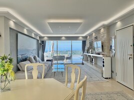 A modern living room with a sofa, dining table, and chairs, a large window with a city view, and a kitchen area with a refrigerator and counter.