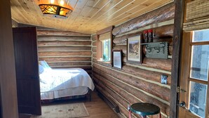 Une chambre rustique en rondins de bois, avec un lit, un tabouret et des tableaux encadrés accrochés au mur.