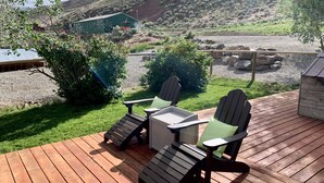 Une terrasse en bois, agrémentée de chaises Adirondack et d’une table, qui offre une vue sur un espace recouvert de gravier et un bâtiment verdoyant, le tout dans un paysage vallonné.