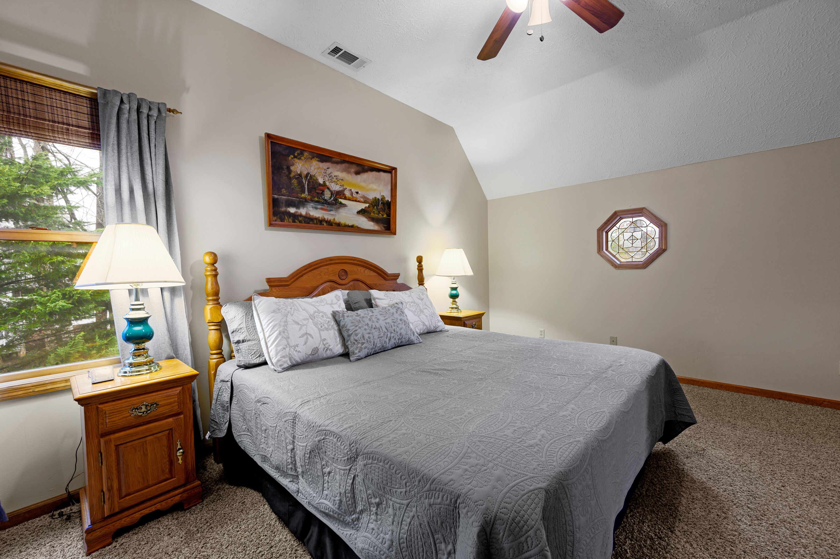 Une chambre à coucher comprenant un lit, deux tables de chevet, une fenêtre avec des rideaux, un ventilateur de plafond, un tableau accroché au mur et un élément décoratif mural.