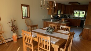 Uma área de refeições com cadeiras e mesa de madeira, uma cozinha com armários de madeira, e uma janela com vista para o verde.