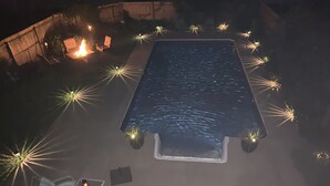 Uma piscina retangular com um deck ao redor, uma pequena mesa com uma cadeira e um lareiro.