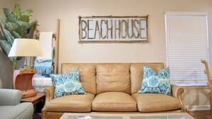 Un soggiorno con un divano in pelle color cuoio, due cuscini decorativi, una targa di legno con la scritta "BEACHHOUSE", una lampada e una pianta.