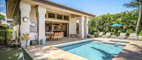 Un espace aménagé au bord de la piscine, avec un patio couvert, du mobilier d’extérieur et une vue sur une maison à colonnes.