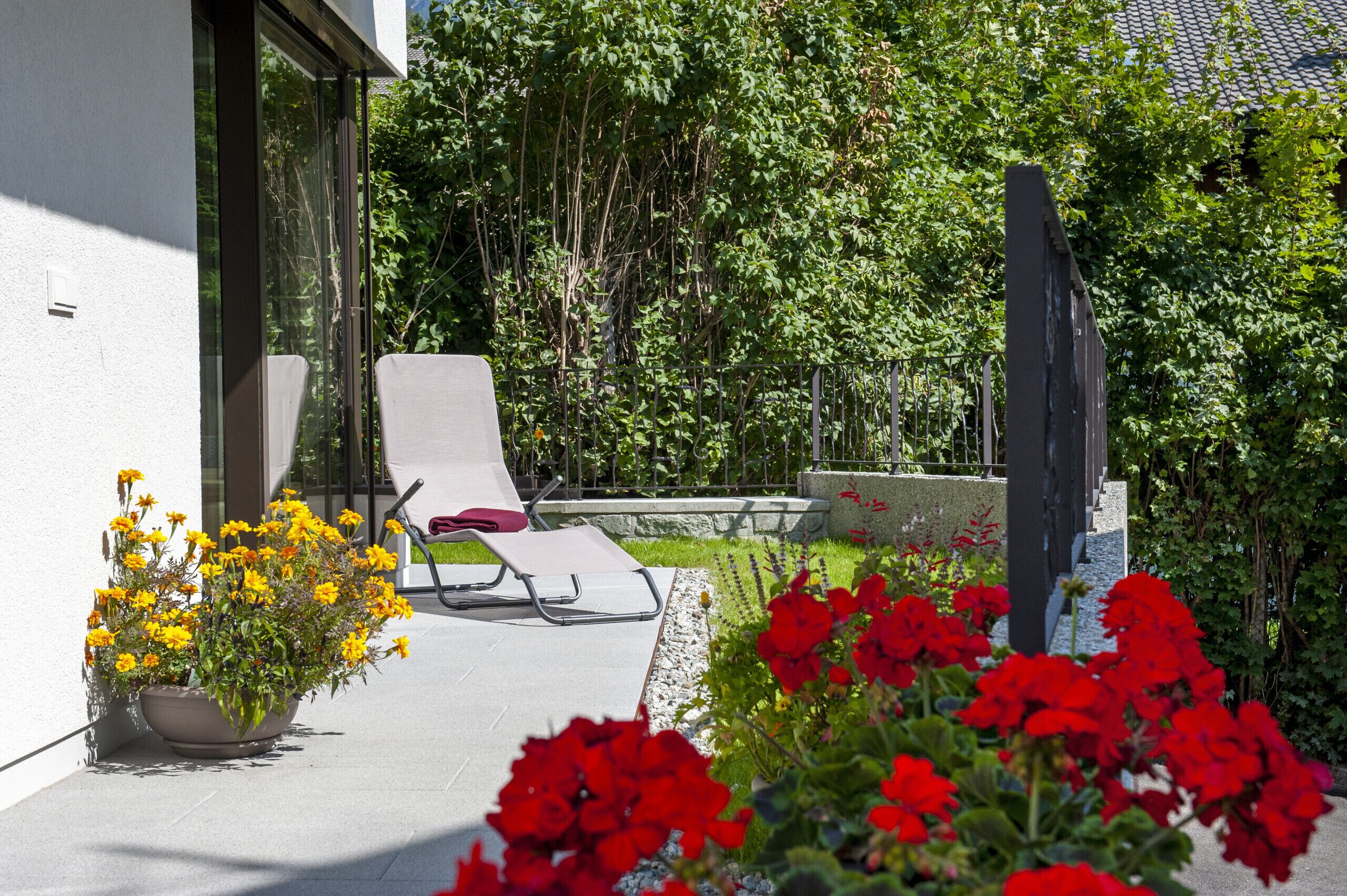 A modern outdoor seating area with a lounge chair and a table, surrounded by potted plants and flowers.