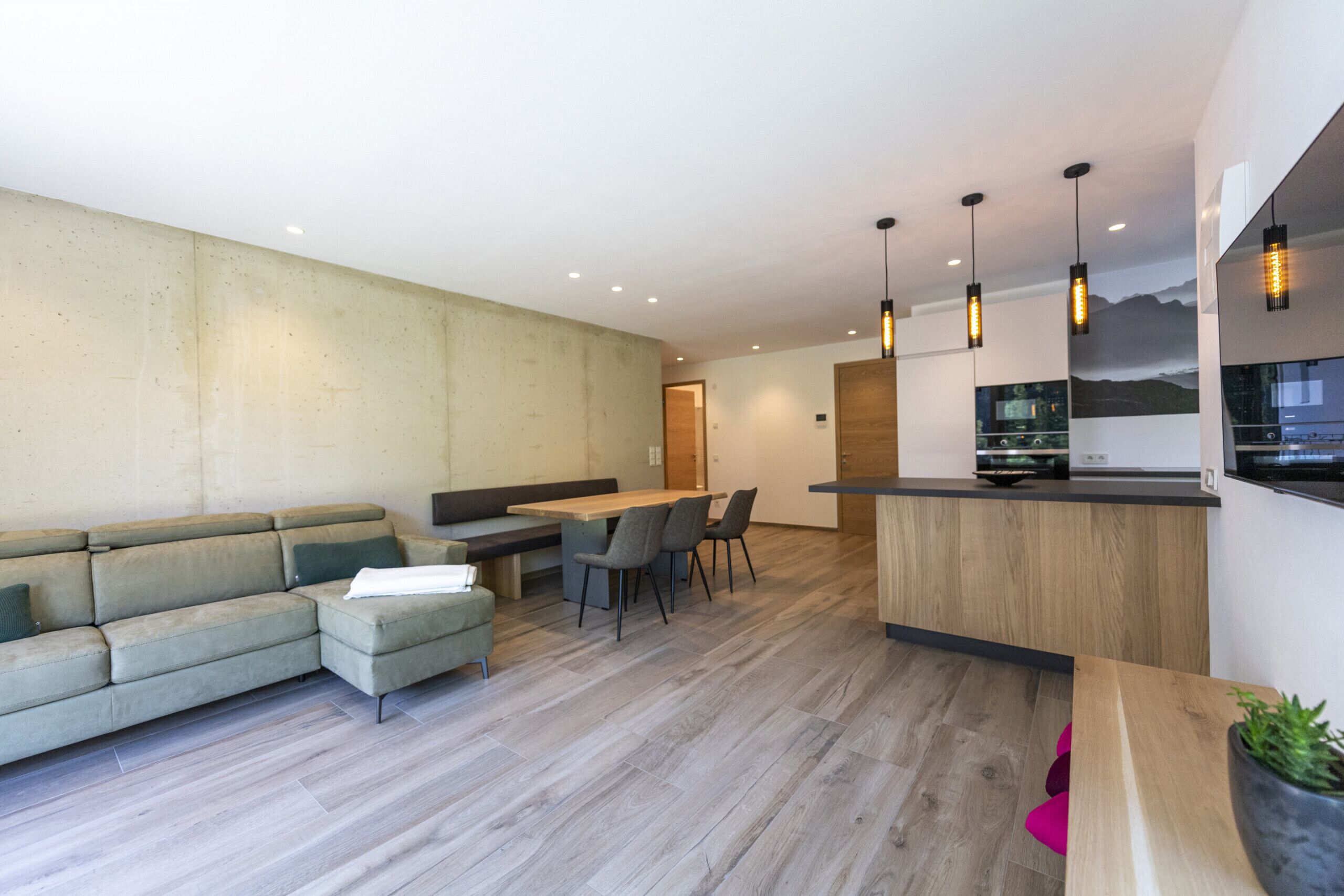 A modern living room with a sofa, dining table, and chairs. There is a kitchen area with a counter and hanging lights.