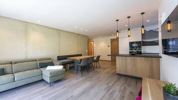 A modern living room with a sofa, dining table, and chairs. There is a kitchen area with a counter and hanging lights.
