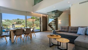 Un salon moderne avec un canapé, une table basse et un coin repas avec des chaises. On aperçoit un escalier au fond.