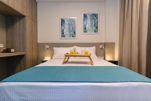 Une chambre d’hôtel comprenant un lit, une table de chevet, une étagère avec quelques objets, deux œuvres d’art murales et une fenêtre avec des rideaux.