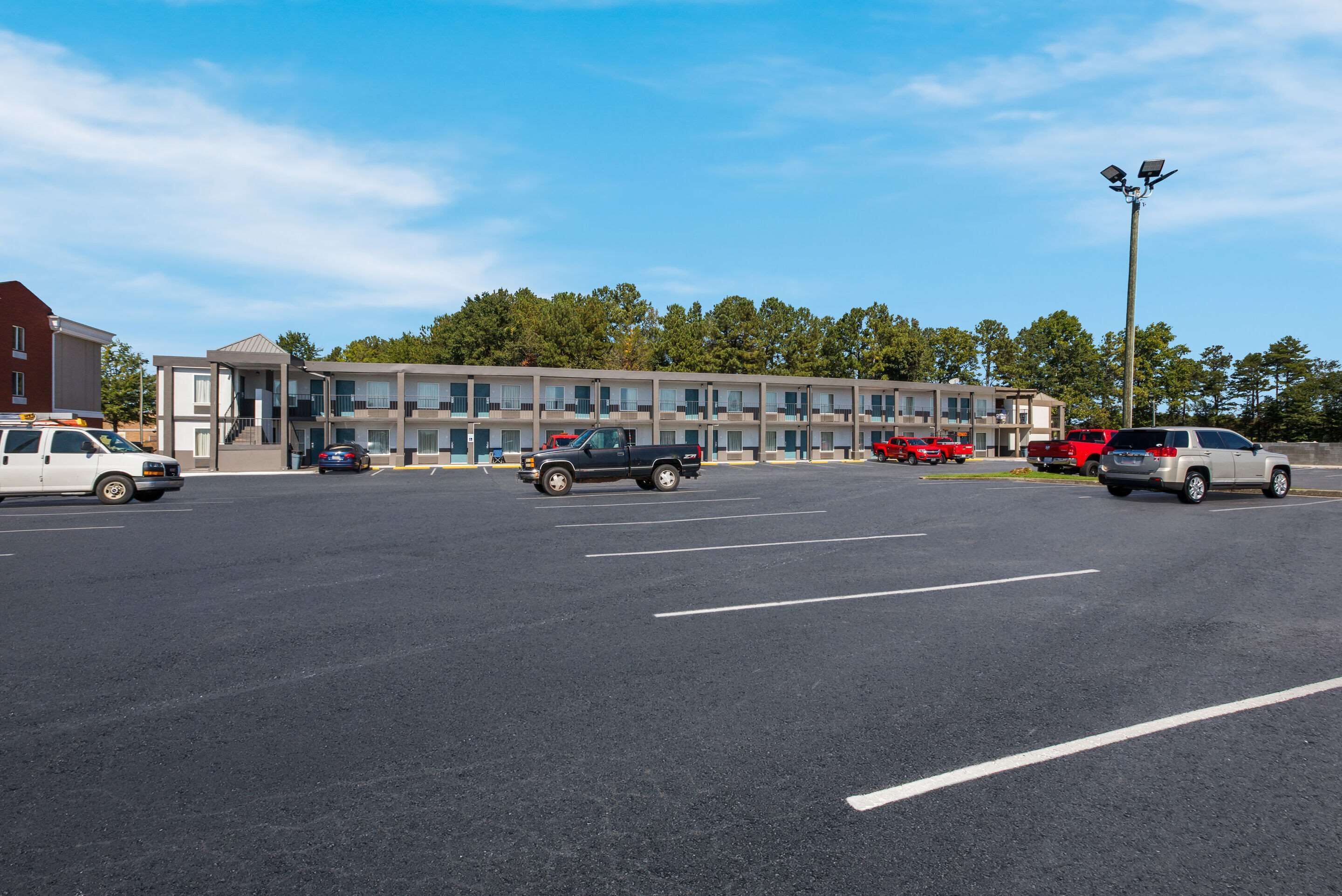 A parking lot with several cars, a building with multiple balconies, and a row of trees in the background.