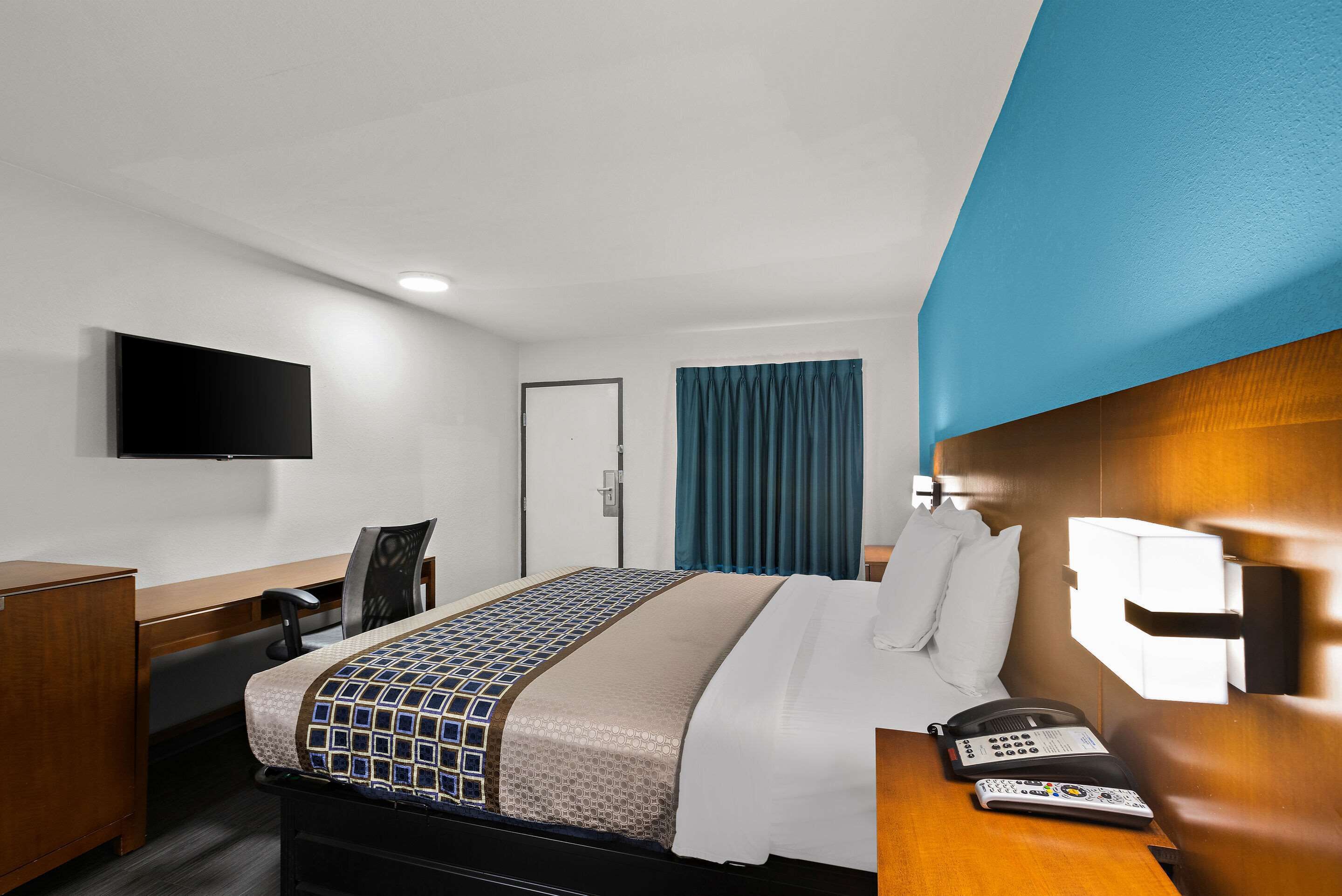 Ein Hotelzimmer mit einem Bett, einem Schreibtisch, einem Sessel, einem Telefon und einem an der Wand befestigten Fernseher.