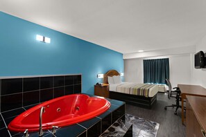 A hotel room with a red jacuzzi, a bed with a wooden headboard, a desk, and a television. - Rodeway Inn Commerce (Commerce)