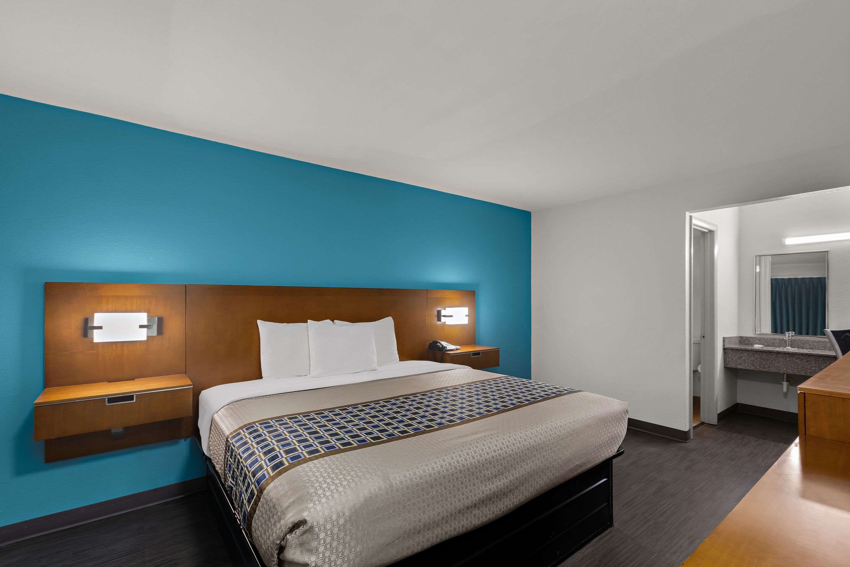 Habitación de hotel con una cama grande, una mesita de noche con lámpara, un baño con espejo y lavamanos, y un escritorio de madera.