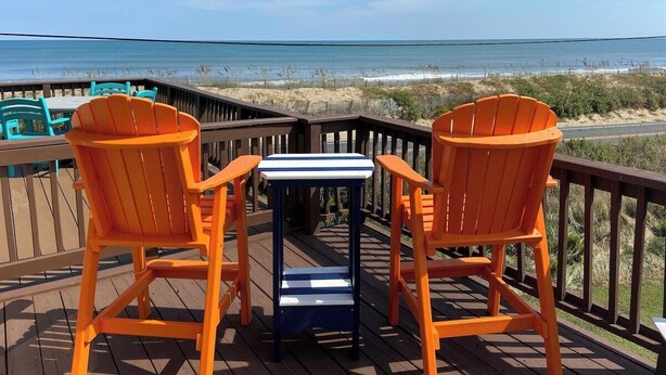 A wooden deck with two orange Adirondack chairs and a small table, overlooking a beach with a fence and vegetation. - Superheat Retreat - Just 75 step to the beach. Breathtaking ocean view. (Kitty Hawk)