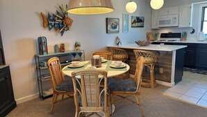 A dining area with a round table, wicker chairs, and a kitchen in the background. - Superheat Retreat - Just 75 step to the beach. Breathtaking ocean view. (Kitty Hawk)
