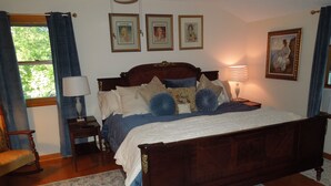 Quarto com cama de madeira, mesa de cabeceira com abajur, uma cadeira e janela com cortinas. Há três quadros na parede acima da cama.