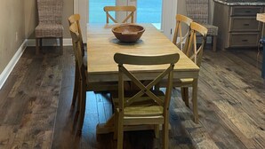 Un coin repas avec une table en bois, quatre chaises, un luminaire central et une vue à travers une porte.