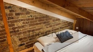 A bedroom with a brick wall, wooden beams, a bed with white bedding, and a patterned pillow.
