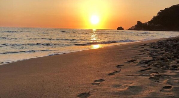 A beach with footprints leading to the water, a sunset, and a cliff in the background.
