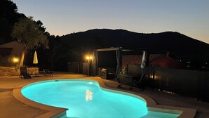 A swimming pool with a curved edge, surrounded by a stone deck. There are lounge chairs and a covered seating area with a table. The setting is outdoors with a view of a hill in the background. - Very cosy villa with swimming pool 20 minutes from Nice (Drap)