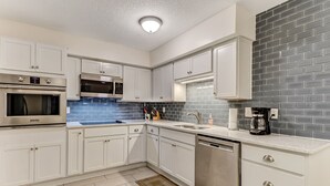 A modern kitchen with white cabinets, stainless steel appliances, a blue backsplash, and a gray brick accent wall. - Beautiful family beach townhome getaway (Fernandina Beach)