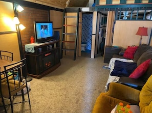 Cozy living space with a television, dining table, and sofa, featuring a ladder and door to a bathroom.