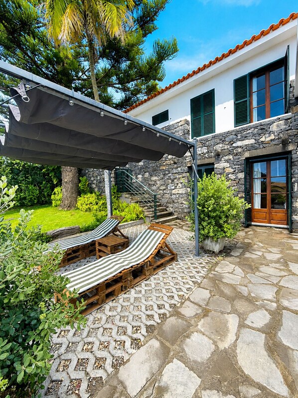 A patio with lounge chairs and a pergola, a stone pathway, and a building with stone walls and a tiled roof.