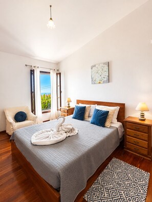 A hotel room with a bed, a chair, a nightstand, a lamp, a window with curtains, and a painting on the wall.