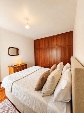 Ein Schlafzimmer mit einem hölzernen Kleiderschrank, einem Nachttisch mit Lampe, einem Bett mit vielen Kissen und einem gemusterten Teppich.