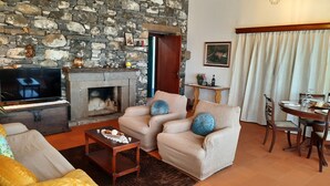 Uma sala de estar com parede de pedra, lareira, televisão, dois poltronas, uma mesa de centro e um espaço de jantar com mesa e cadeiras.
