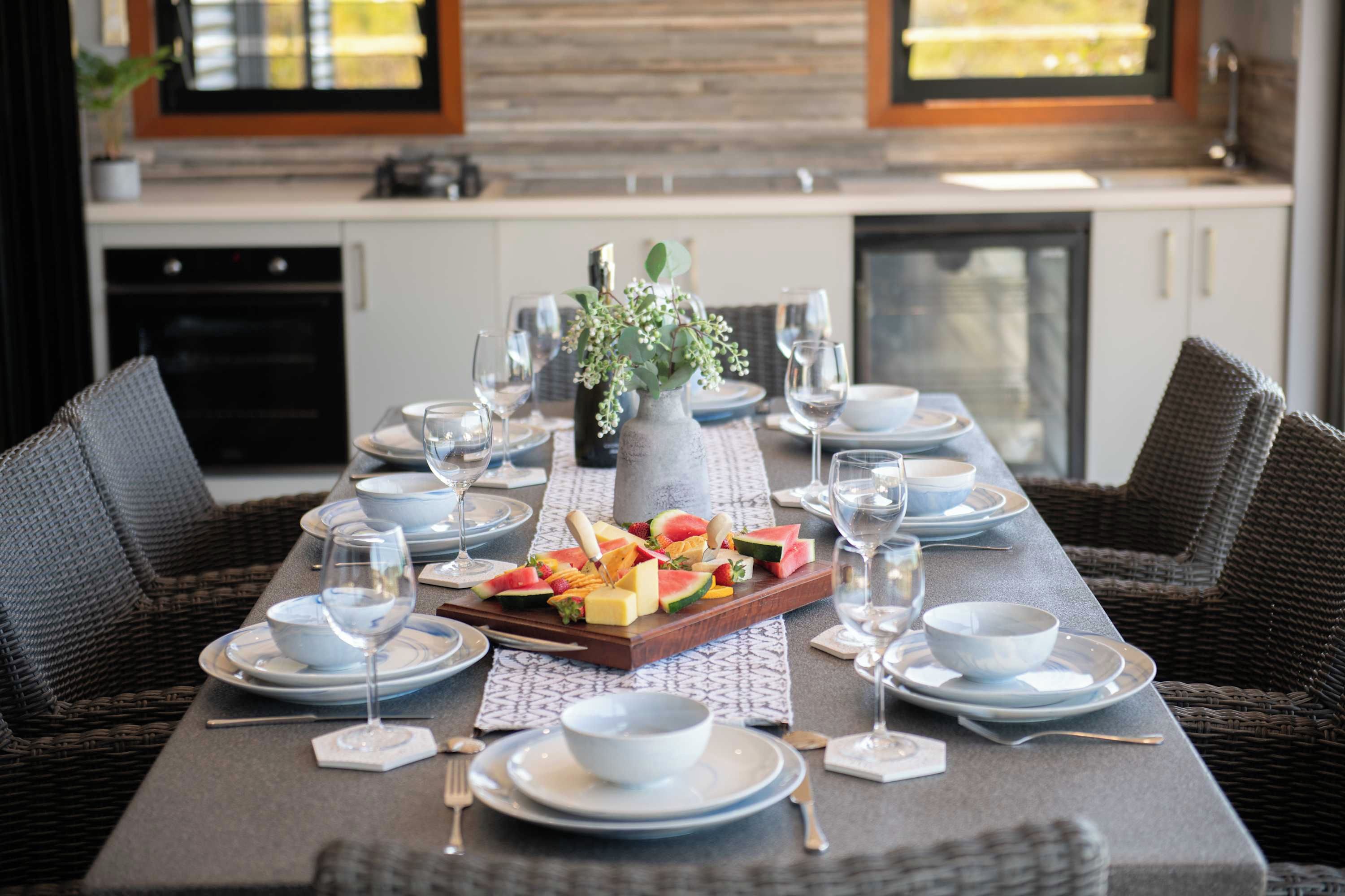 A dining table set with plates, bowls, glasses, and a fruit platter, with a kitchen in the background.