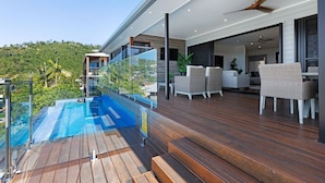 A modern outdoor deck with a glass balustrade overlooking a pool, wooden flooring, and outdoor furniture.
