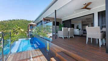 A modern outdoor deck with a glass balustrade overlooking a pool, wooden flooring, and outdoor furniture.