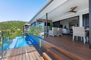 A modern outdoor deck with a glass balustrade overlooking a pool, wooden flooring, and outdoor furniture. - Lux On Nara (Airlie Beach)