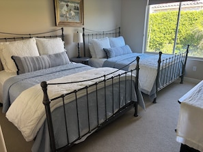 A bedroom with two beds, a bedside table with a lamp, and a window with a view of greenery. - Sun Drenched Tuscan home! 4 Bedrooms 4.5 Bathrooms, 2 Primary Suites, 2 Bedrooms (La Quinta)