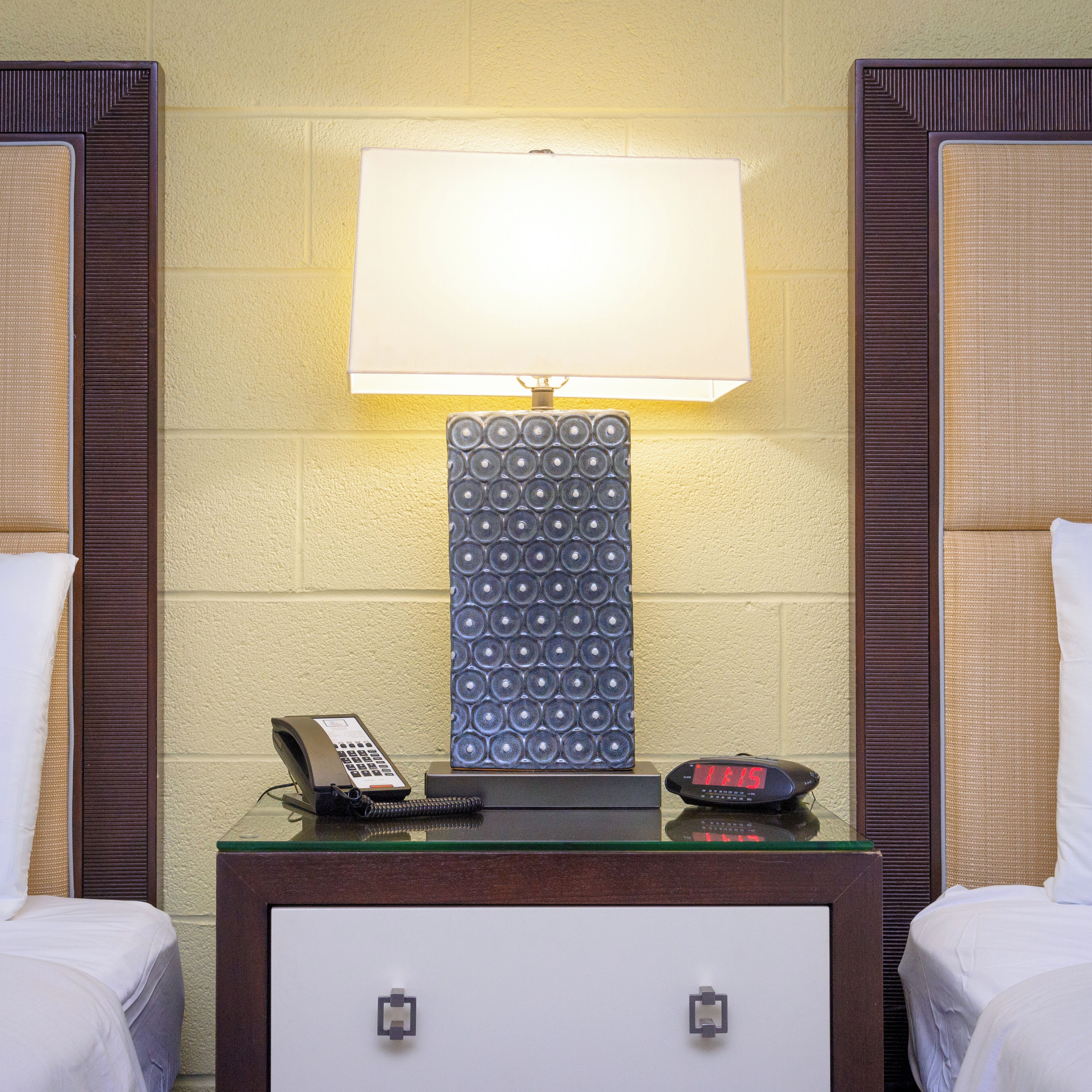 A hotel room with a bedside table, a lamp, a phone, and a digital clock.